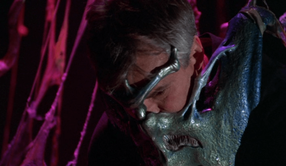 Man sieht Jack O'Neill mit einer Alienmaske über dem Gesicht, er scheint zu schlafen. Das Standbild ist aus der Szene, in der er so tut, als ob er noch in Ruhe an der Decke hängt.