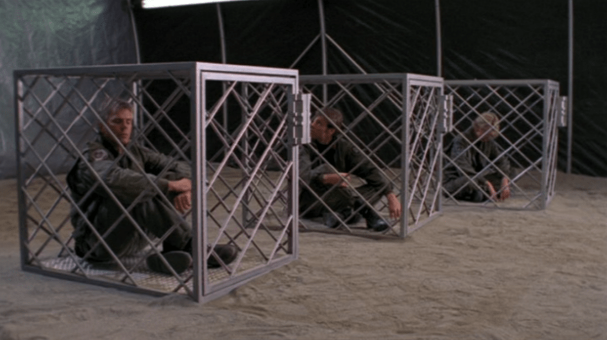 Jack, Sam und Daniel sitzen in Käfigen, auf einem Sandboden in einem Zelt.